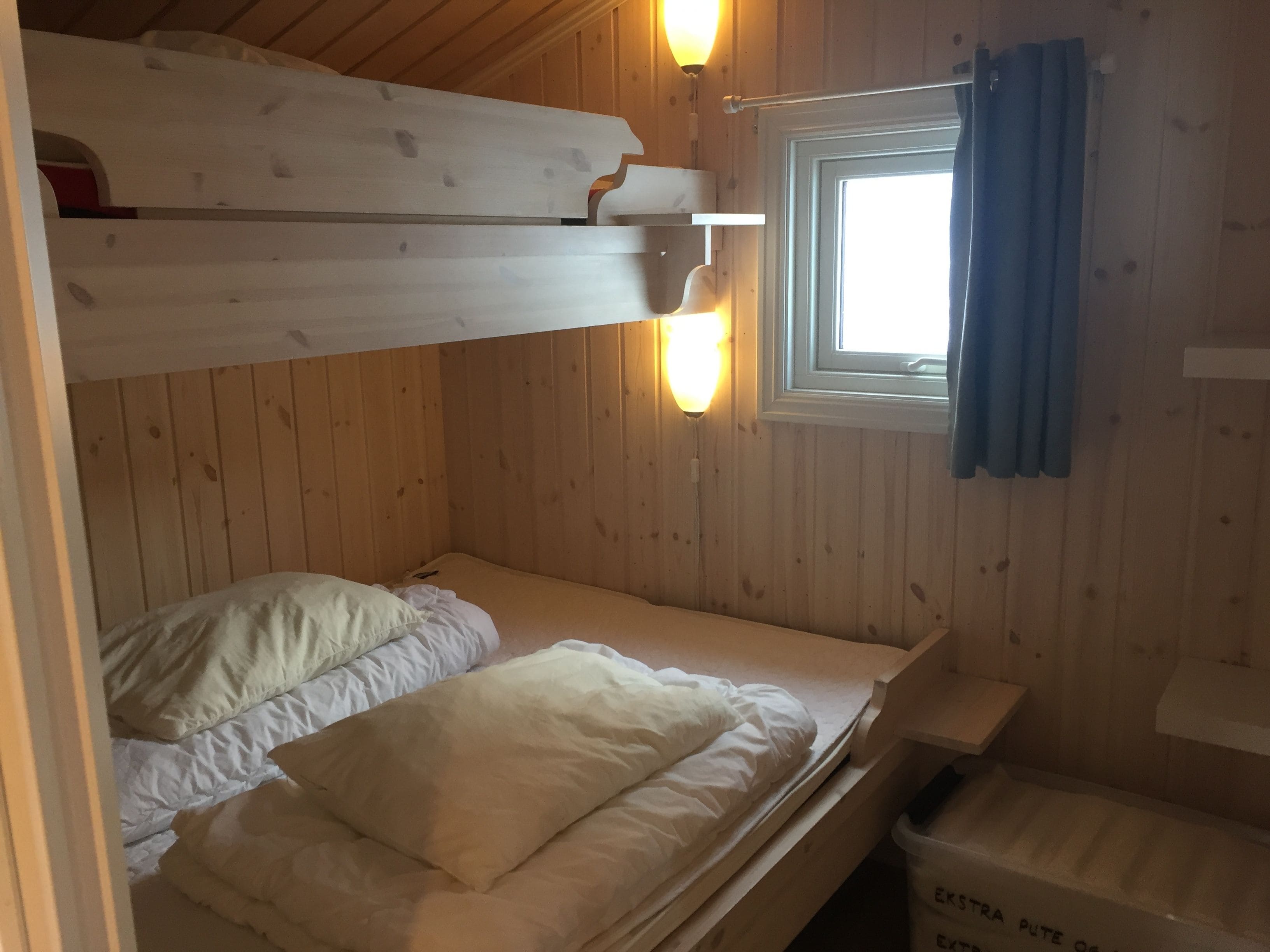 A bunk bed room with wooden walls, a window with curtains, and a small table.