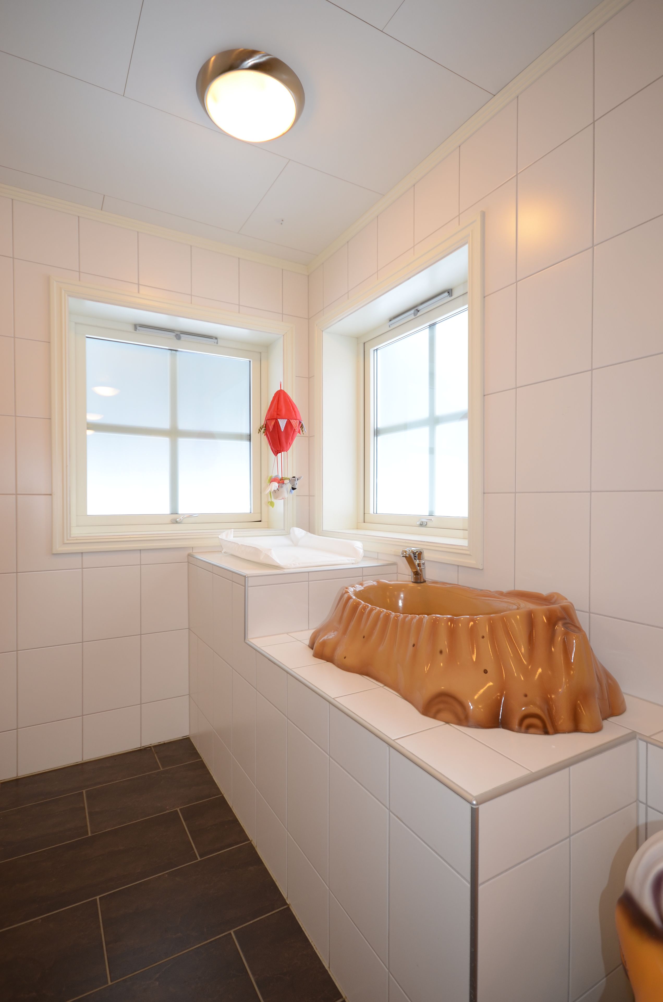 A bathroom with two windows, a white sink, a unique basin, and a red hanging decoration.