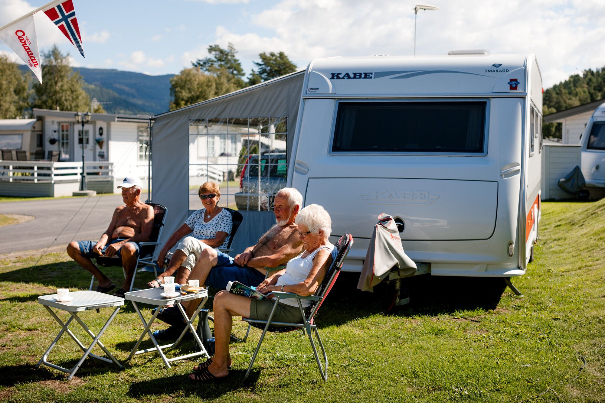 A caravan with a KABE logo, parked on grass with people sitting on chairs.