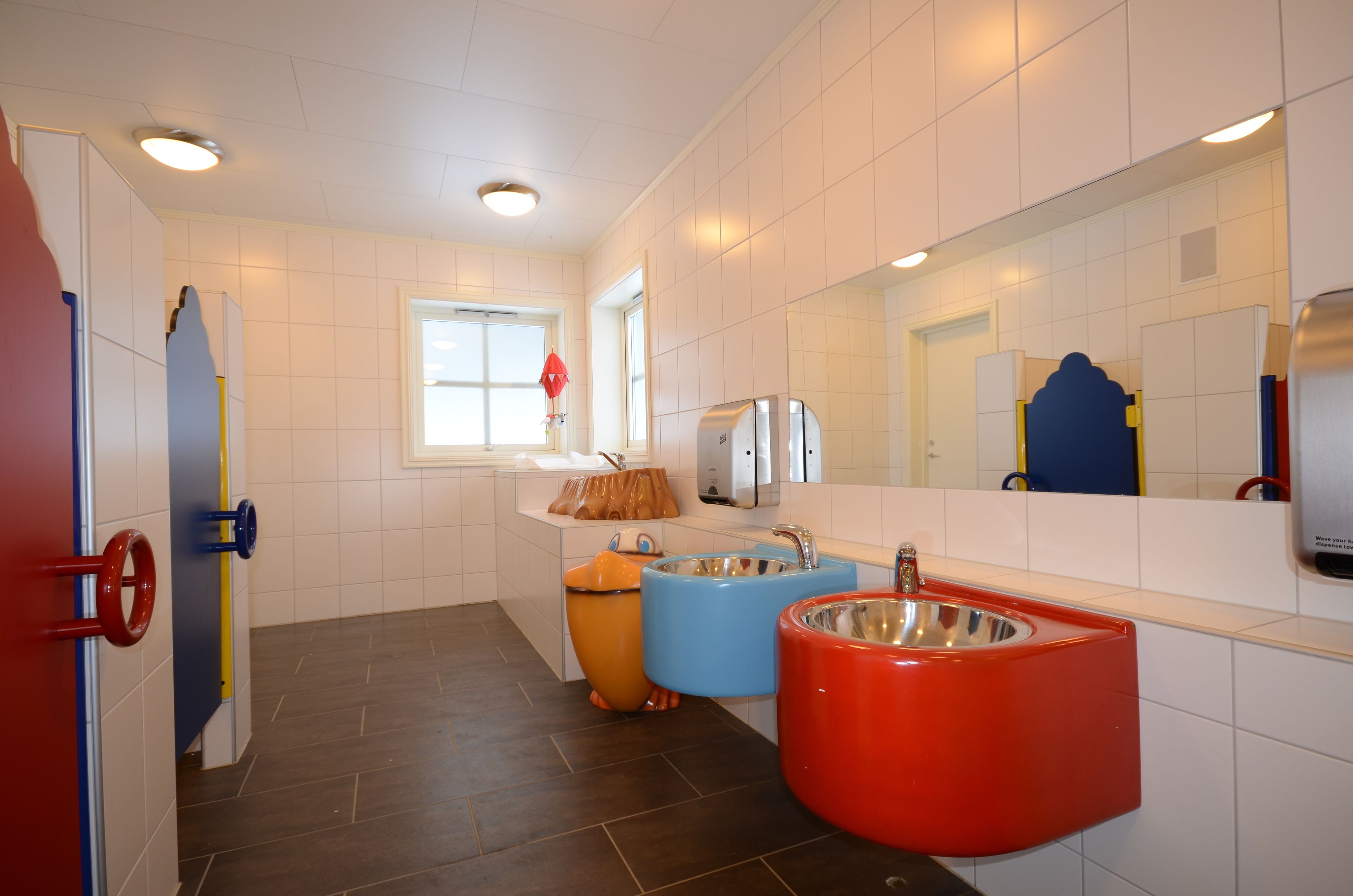 A bathroom with white tiled walls, a red door, and three sinks in blue, orange, and red.