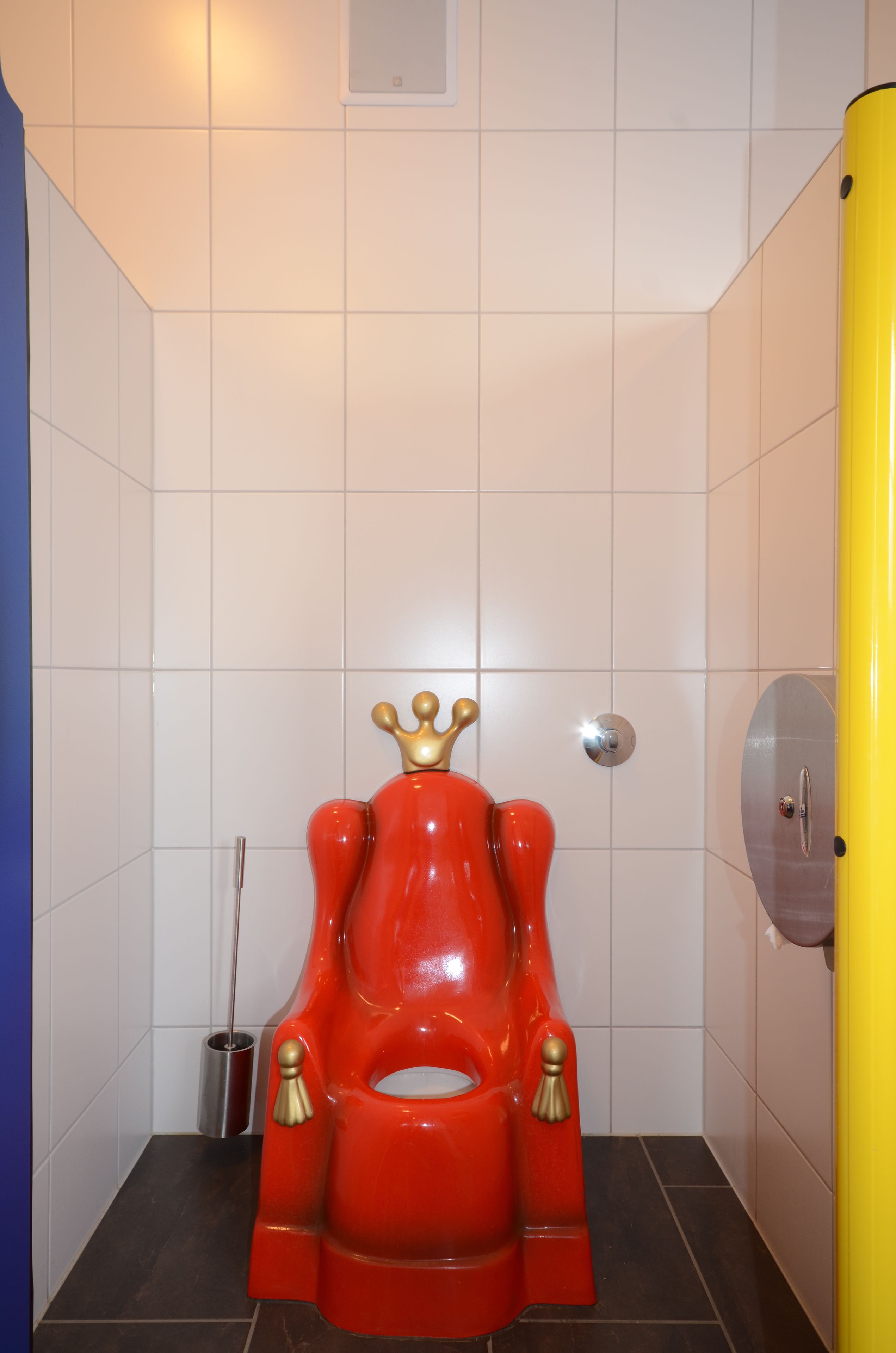 Une salle de bain avec des toilettes dont le couvercle rouge ressemble à un trône, des murs recouverts de carreaux blancs, une porte jaune et une poubelle en métal.