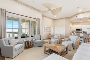 A spacious living room with a large window, a ceiling fan, and a kitchen area. - 303W ~ Captivating 3BR/3BA condo with a Gulf view (Gulf Shores)