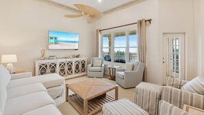 A spacious living room with a large TV, comfortable seating, a ceiling fan, and a view of the ocean. - 303W ~ Captivating 3BR/3BA condo with a Gulf view (Gulf Shores)