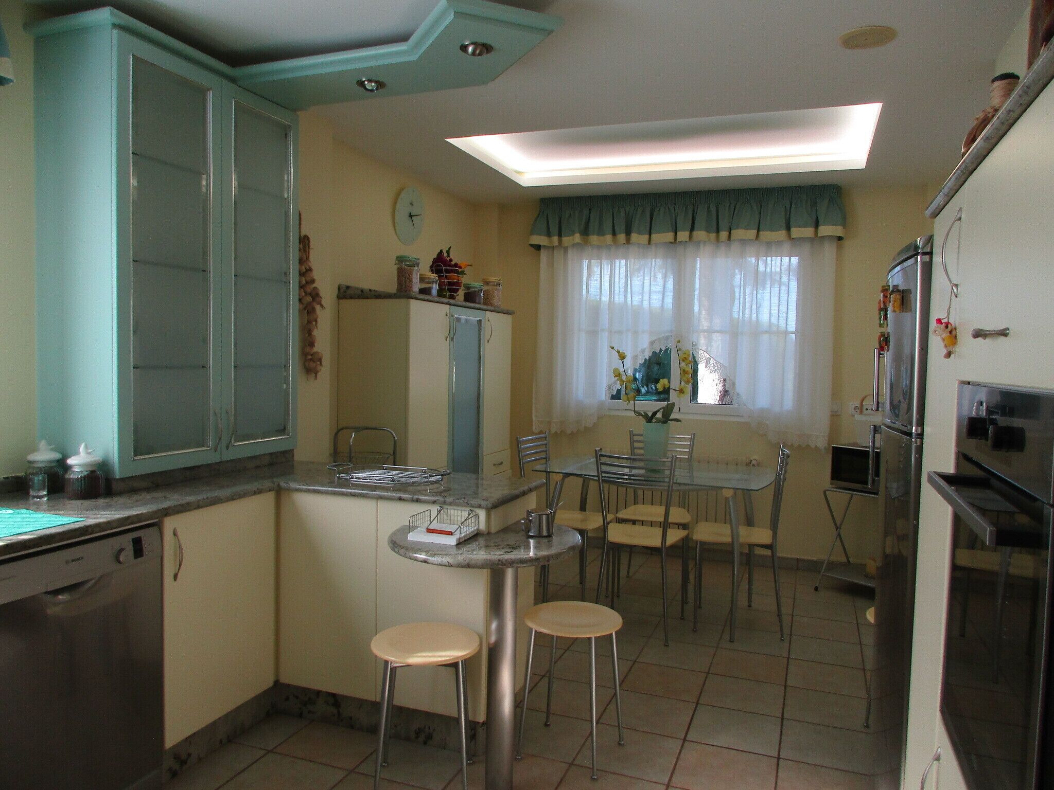 A compact kitchen with a dining area, a clock, and a window with sheer curtains.