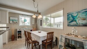 A dining area with a white table, wooden chairs, and a chandelier. There is a window with blinds and a painting of a blue starfish on the wall.