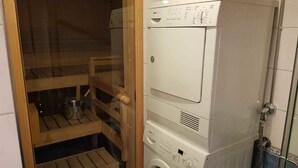 Ett kompakt tvättrum med en tvättmaskin och torktumlare placerade ovanpå varandra, ett träställ för att torka kläder och en liten bänk.