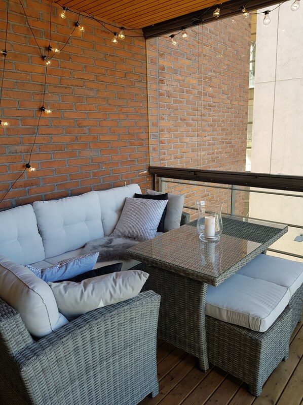 A wicker sofa with cushions, a glass-top table, and string lights against a brick wall.