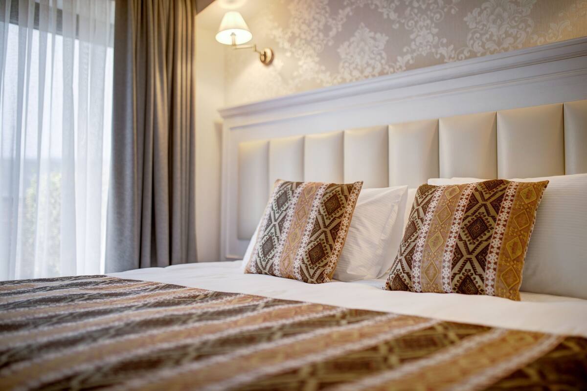 A neatly made bed with patterned bedding, a decorative wall lamp, and a window with sheer curtains.