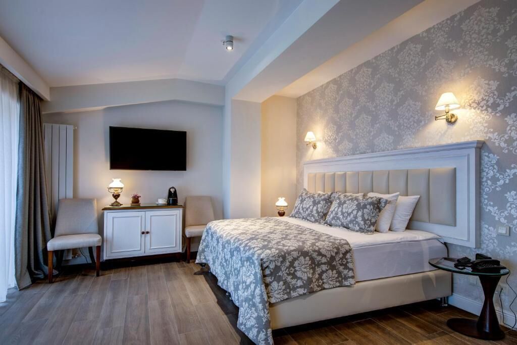 A modern hotel room with a large bed, a flat-screen TV mounted on the wall, a desk with a lamp, and a chair.