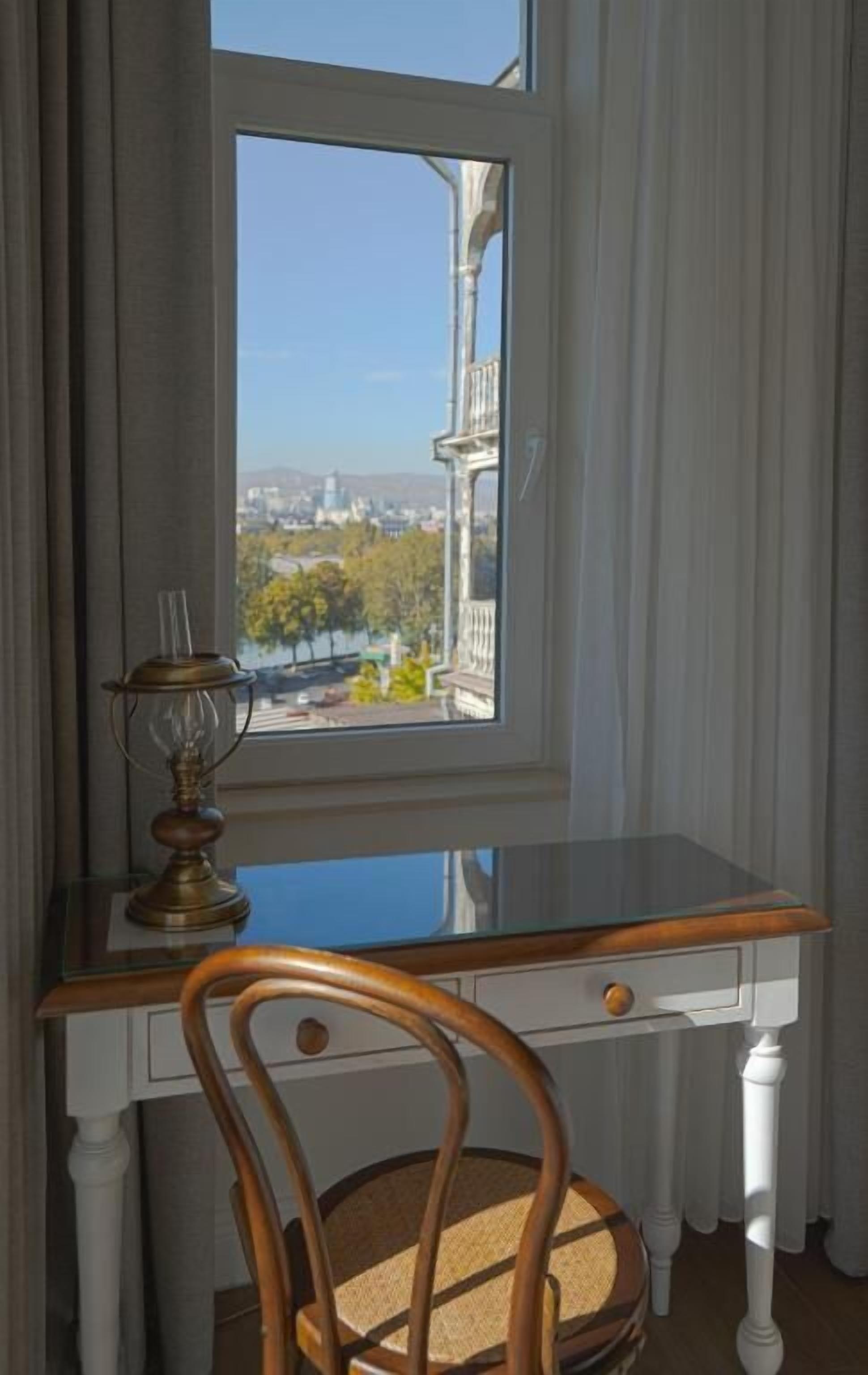 A room with a desk and chair, a window with a view of a cityscape, and a lamp on the desk.