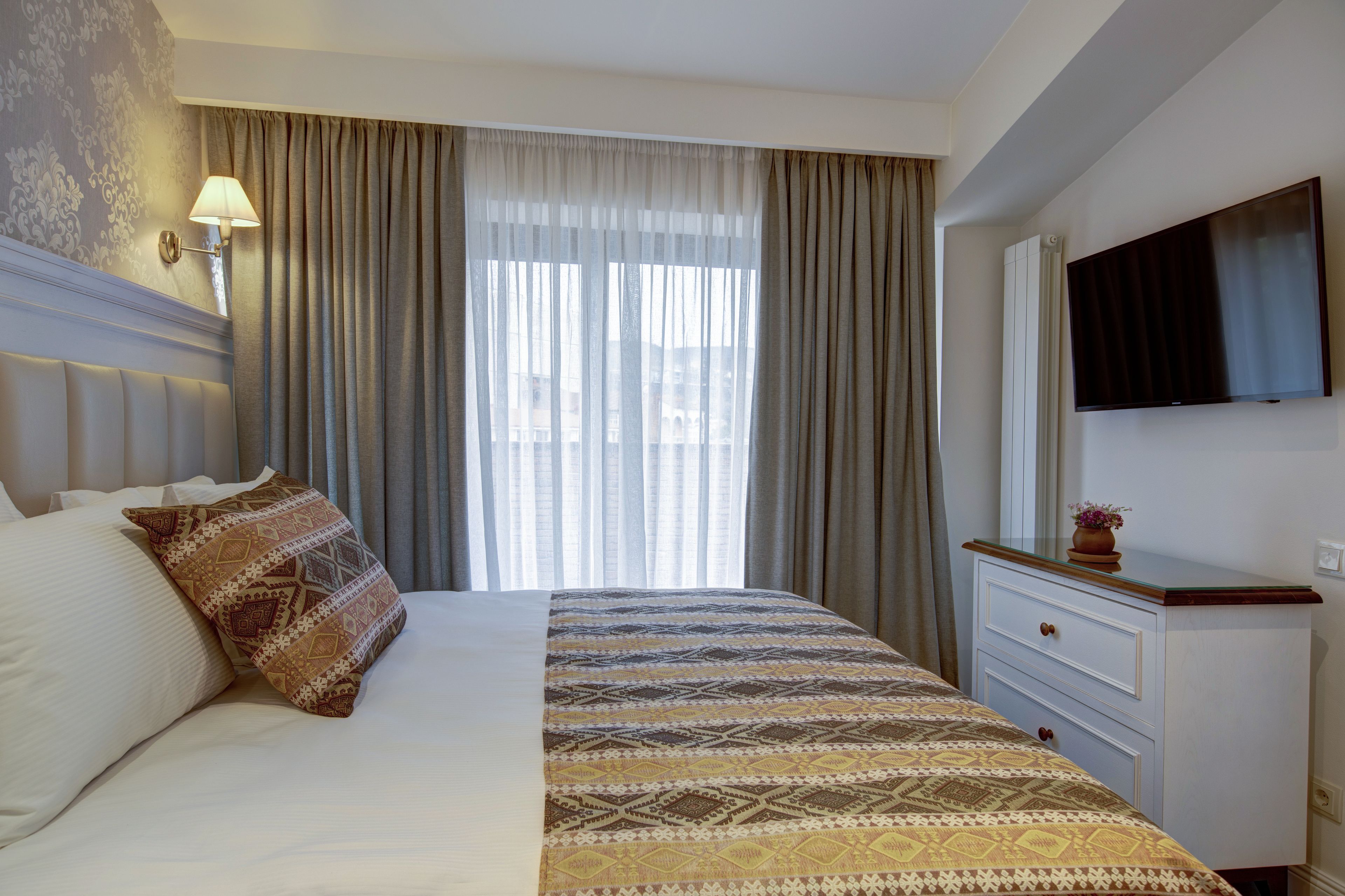 Una habitación de hotel con una cama grande, una mesita de noche con lámpara, un televisor montado en la pared y una ventana con cortinas.