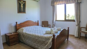 A bedroom with a wooden bed, a chair, a nightstand, a window with curtains, and a crucifix on the wall. - Cozy home in Maroni-Larnaca (Maroni-Larnaca)