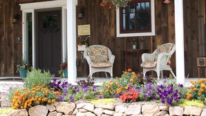 Een houten huis met een veranda, twee rieten stoelen en een verscheidenheid aan kleurrijke bloemen.