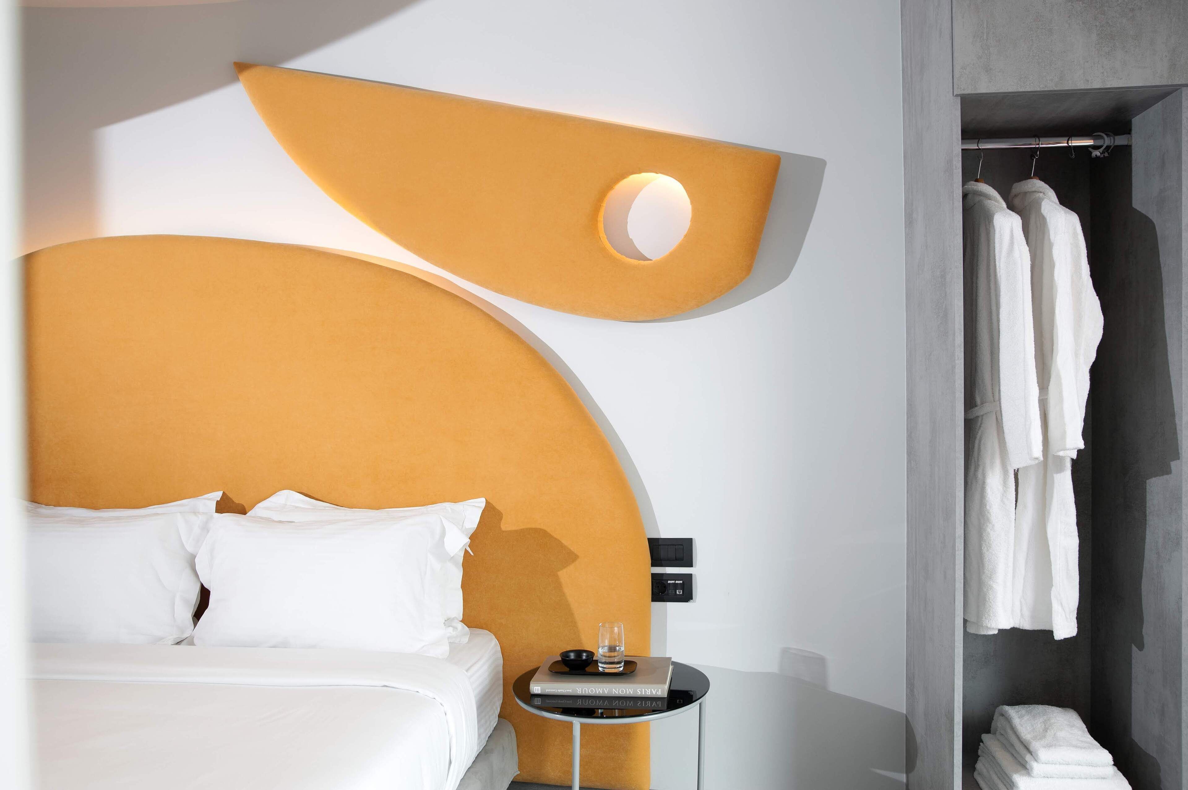 Ein modernes Hotelzimmer mit einem großen Bett, einem Nachttisch mit Buch und Glas sowie einem Kleiderschrank mit Bademänteln.