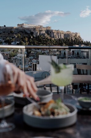 A dining table with dishes and a drink, overlooking ancient ruins and a hill. - Athens Utopia Ermou (Athens)
