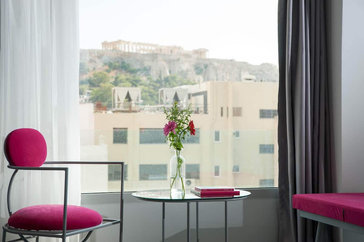Habitación con vista a un edificio histórico, una silla con cojín, una mesita con un florero y libros, y una cama con cubre color rosa.