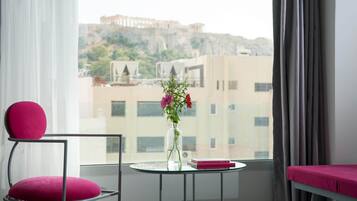 A room with a view of a historic building, a chair with a cushion, a small table with a vase and books, and a bed with a pink cover.