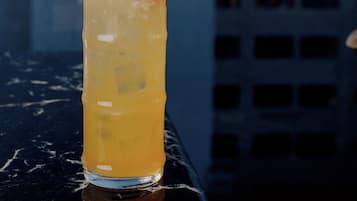A tall glass with a yellow beverage, garnished with a slice of fruit and a red straw, placed on a reflective surface with a cityscape in the background.