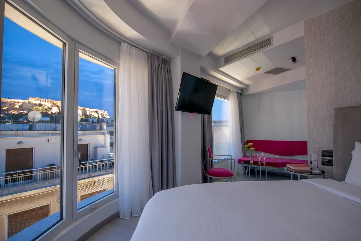 Habitación de hotel con una cama grande, un televisor de pantalla plana en la pared, un balcón con vista a la ciudad y una pequeña zona de estar con un sofá rosa y una mesa.
