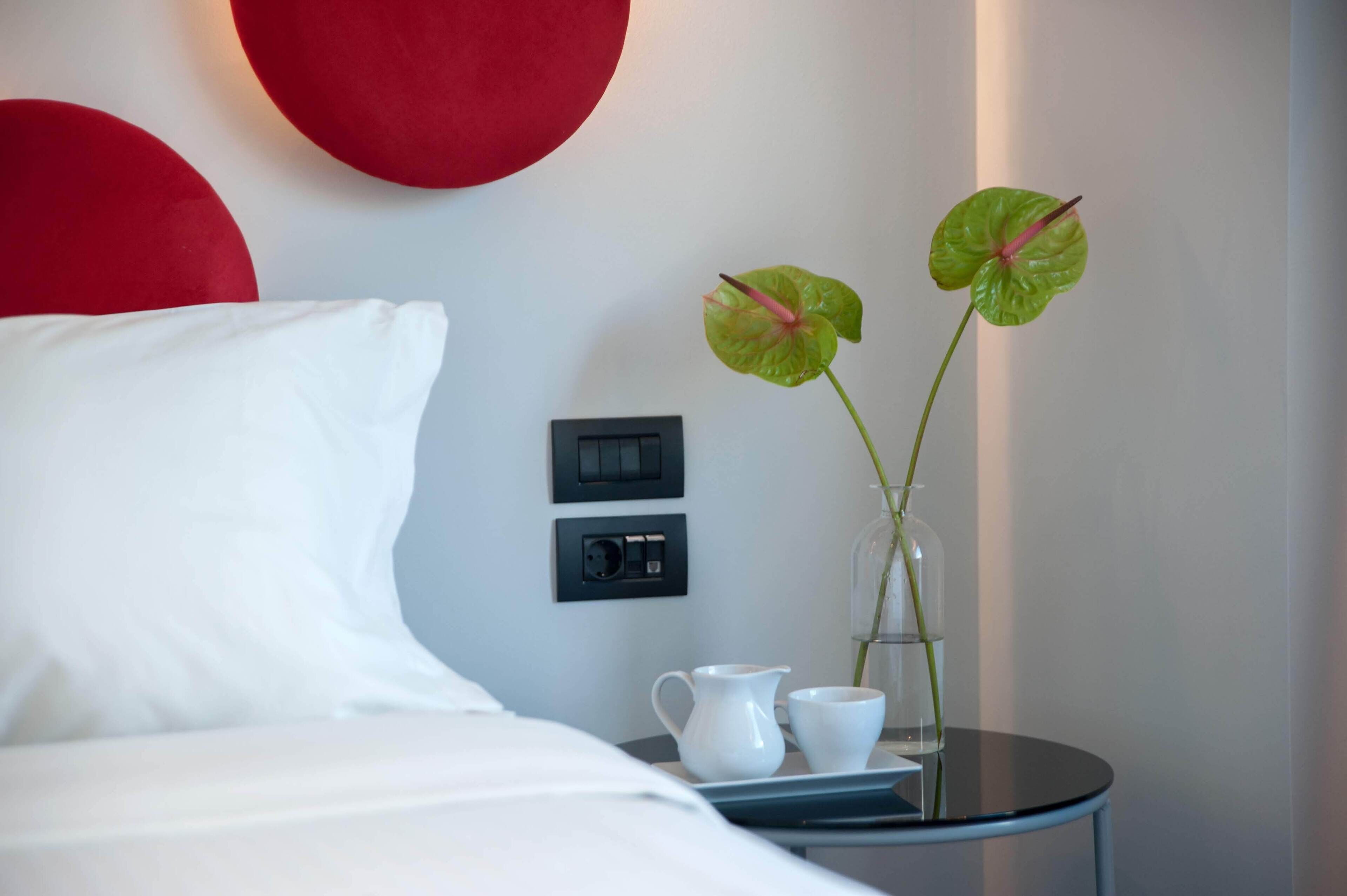 Quarto de hotel com cama, mesa de cabeceira com um vaso de flores, jarra e duas chávenas.