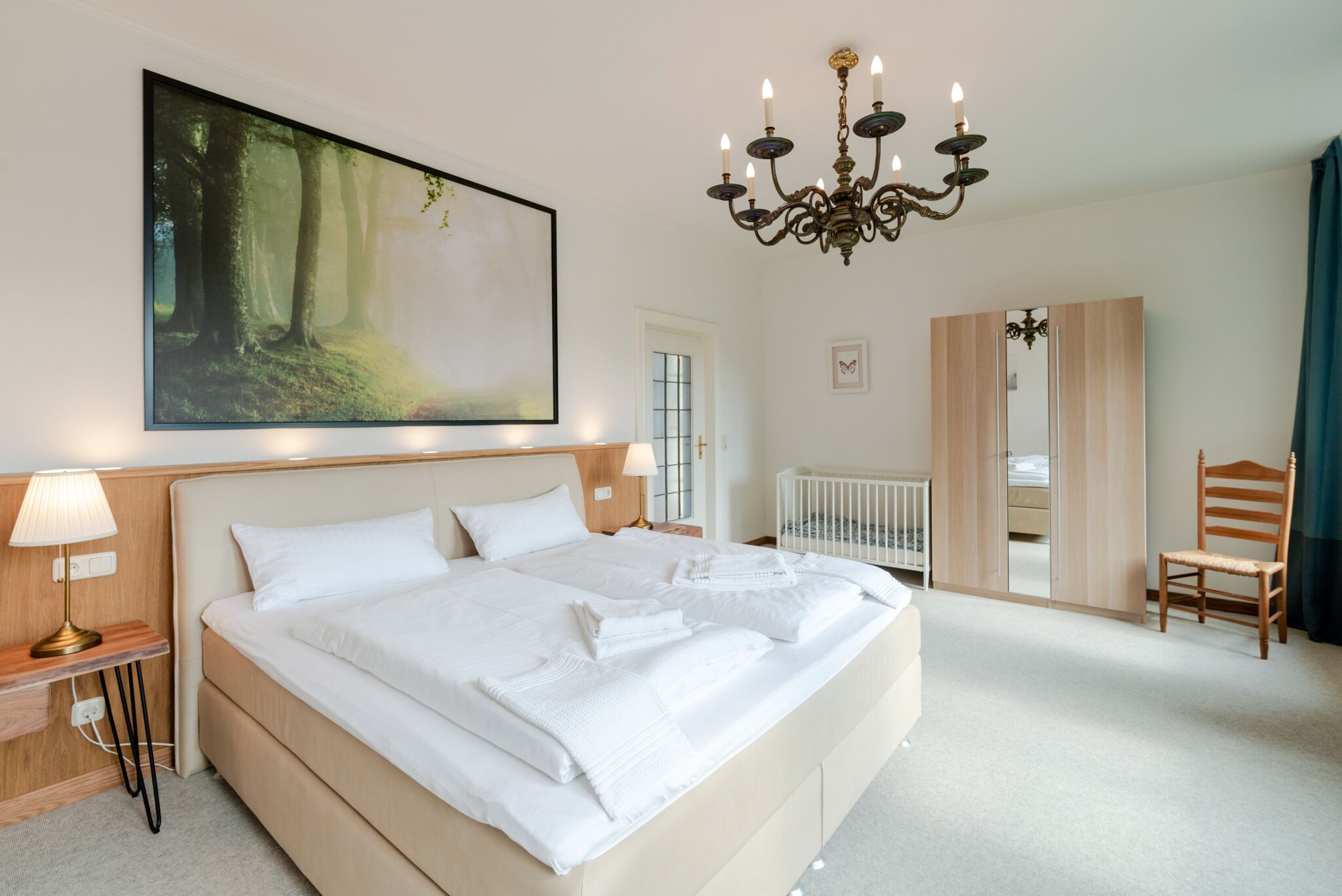 A bedroom with a large bed, a chandelier, a painting of a forest, a crib, and a wardrobe.