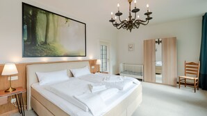 Un dormitorio con una cama grande, una araña, un cuadro de un bosque, una cuna y un armario.
