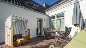 Un patio con terraza de madera, una silla de playa a rayas, una mesa con sillas y un ventanal con vistas a la terraza.