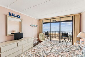 Una habitación de hotel con cama, escritorio, silla, televisor y un amplio ventanal con vista a la playa, además de un letrero que dice 'RELAX'.