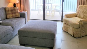A hotel room with a sofa, ottoman, armchair, and a view of the sea through a sliding glass door. - XPLORIE ACTIVITIES & LINENS INCLUDED*!  Direct Oceanfront, 2BR/den, 2BA Plaza condo with Indoor/Outdoor Pools & Tiki Bar! (Ocean City)