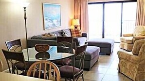 A hotel room with a dining table, chairs, a sofa, a coffee table, and a large window with curtains. - XPLORIE ACTIVITIES & LINENS INCLUDED*!  Direct Oceanfront, 2BR/den, 2BA Plaza condo with Indoor/Outdoor Pools & Tiki Bar! (Ocean City)