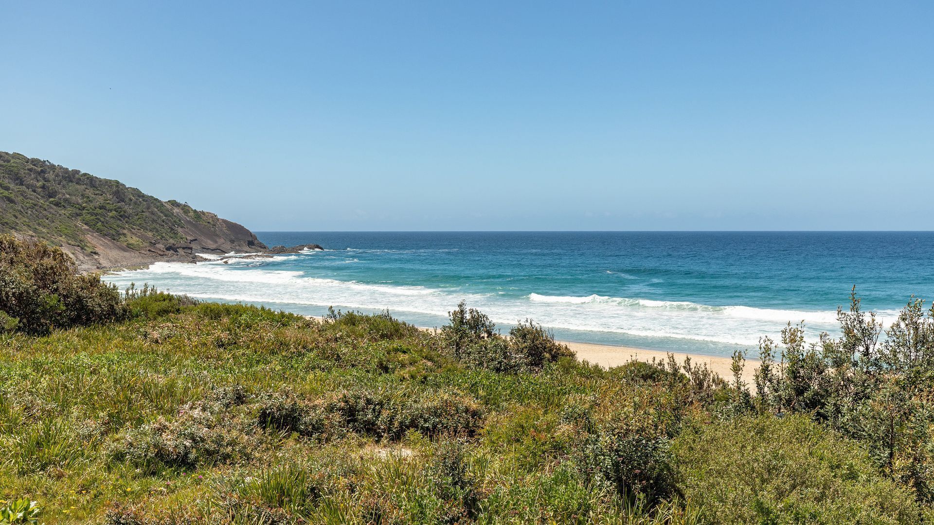 A coastal landscape with a beach, ocean waves, and vegetation.