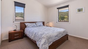 A bedroom with a wooden bed, two bedside tables, a nightstand, and two windows with views of greenery.