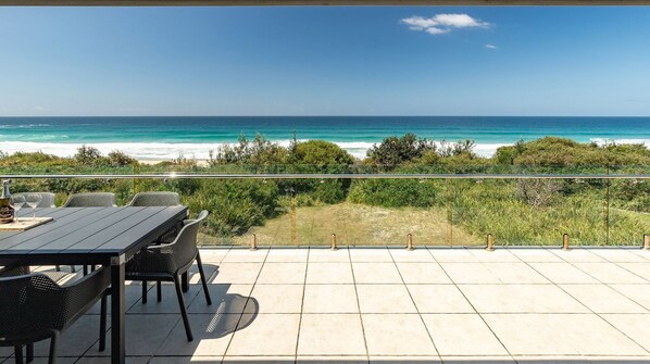 A terrace with a table and chairs overlooking a beach and ocean.