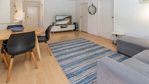 A modern living room with a grey sofa, a dining table with two chairs, a TV on a stand, and a blue striped rug. - Bella Villa Manyana 19 great location short stroll to the beach and local shops (Blueys Beach)