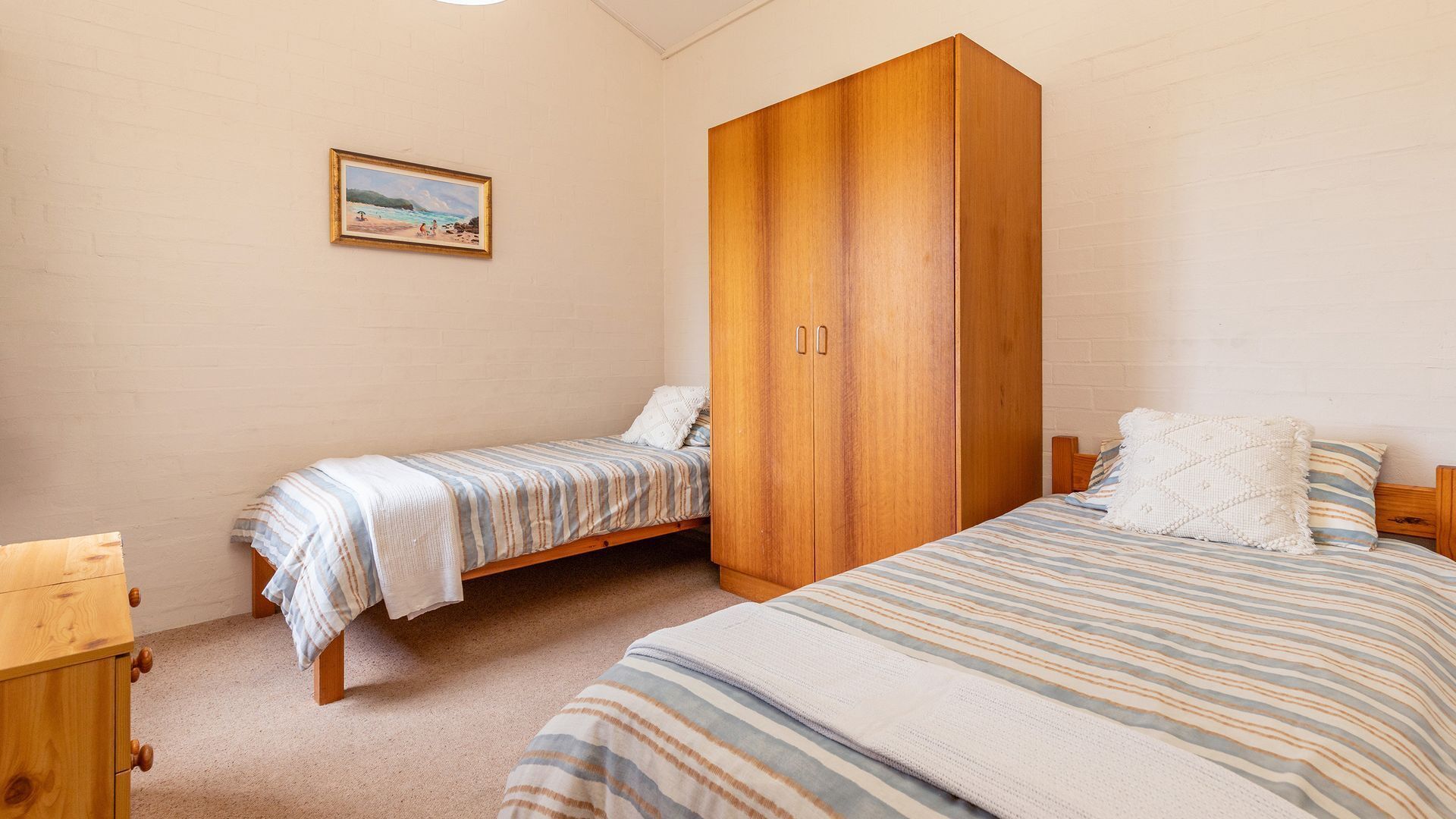A bedroom with two single beds, a wooden wardrobe, a small bedside table, and a framed picture on the wall.