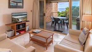 A modern living room with a flat-screen TV, a coffee table, and a sofa. There is a balcony with a table and chairs, and a view of the ocean.