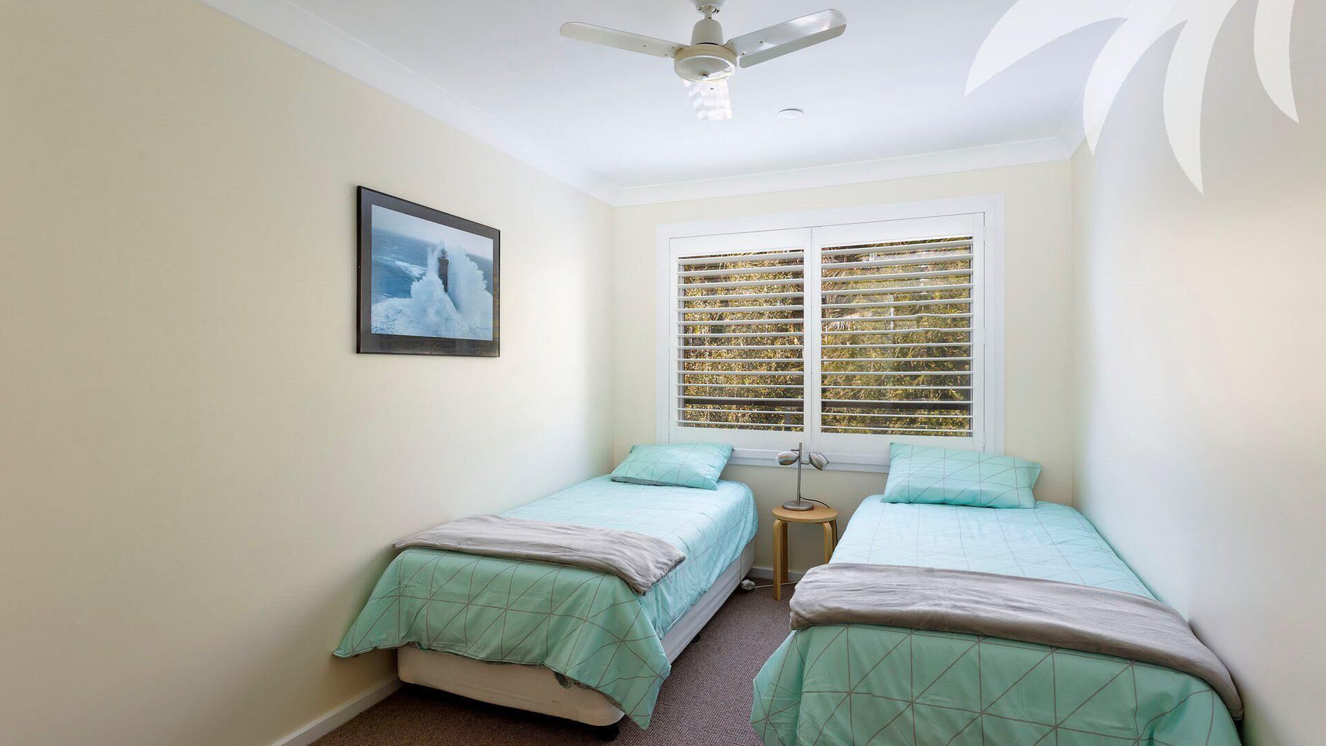 A small, well-lit bedroom with two single beds, a ceiling fan, a window with blinds, and a framed picture on the wall.