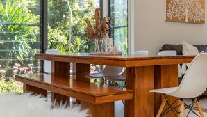 A wooden dining table with a vase of dried plants and a candle, set in a room with large windows and a view of greenery. - Boom 8 - Boomerang Beach (Boomerang Beach)