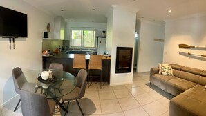 A modern living room with a glass dining table, a sofa, and a kitchen area with a TV, a fireplace, and a window. - Aqua Apartment #2 - Pacific Palms Holidays (Blueys Beach)