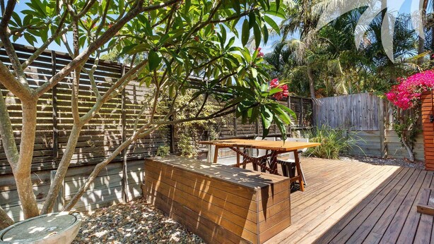 A wooden deck with a table and benches, surrounded by a wooden fence and lush greenery. - Aqua Apartment #2 - Pacific Palms Holidays (Blueys Beach)