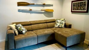 A brown sectional sofa with two decorative pillows, a wall-mounted wooden paddle, and a framed picture. - Aqua Apartment #2 - Pacific Palms Holidays (Blueys Beach)