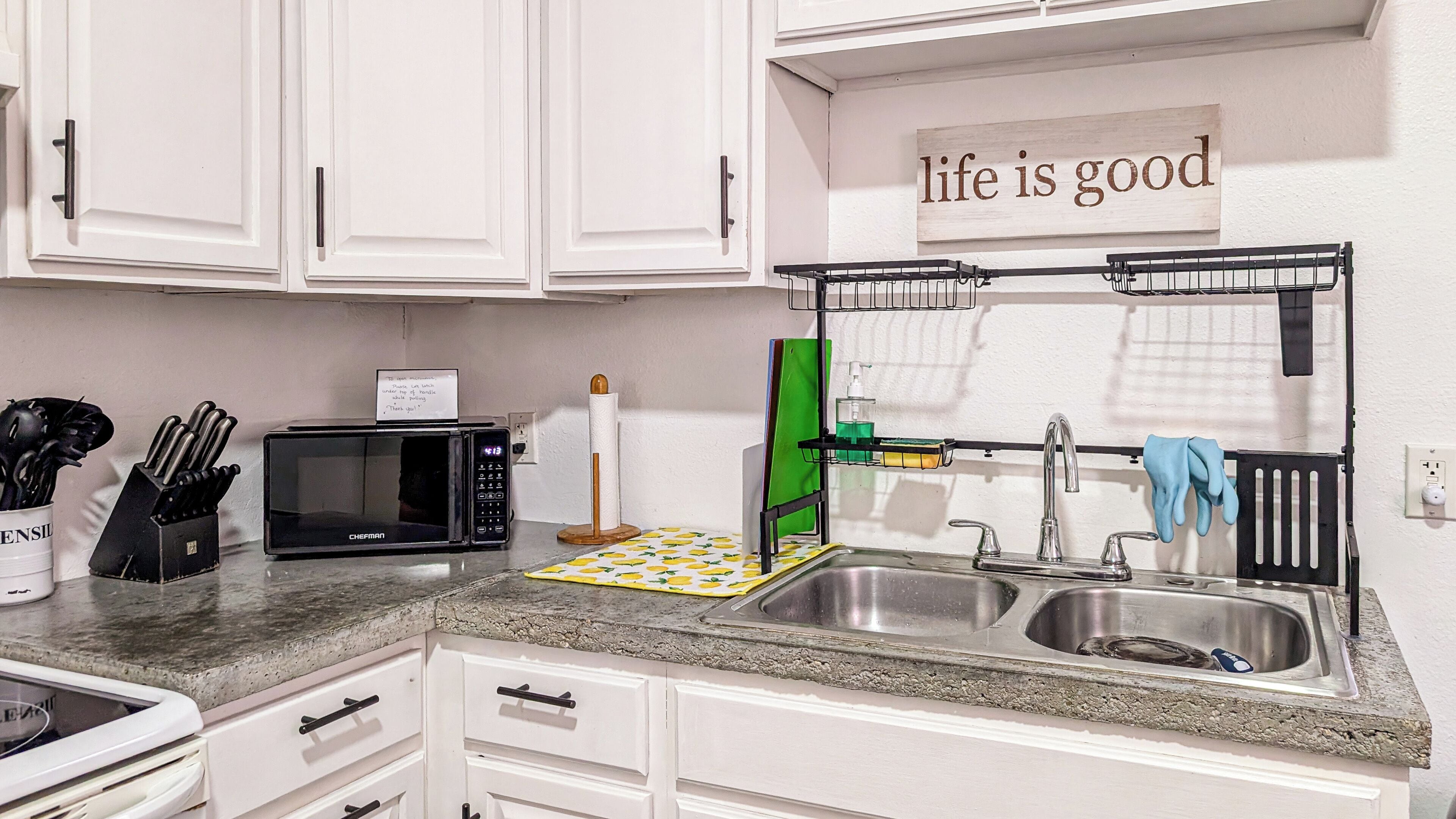 A kitchen with white cabinets, a microwave, a sink with a faucet, and a sign that says 'life is good'.