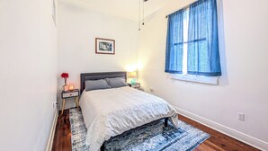 A bedroom with a bed, a nightstand, a lamp, a window with curtains, and a picture on the wall. - Funky Home Away from Home  (New Orleans)
