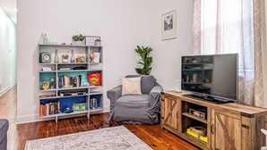 A living room with a grey sofa, a wooden entertainment unit, a bookshelf with various items, and a television. - Funky Home Away from Home  (New Orleans)