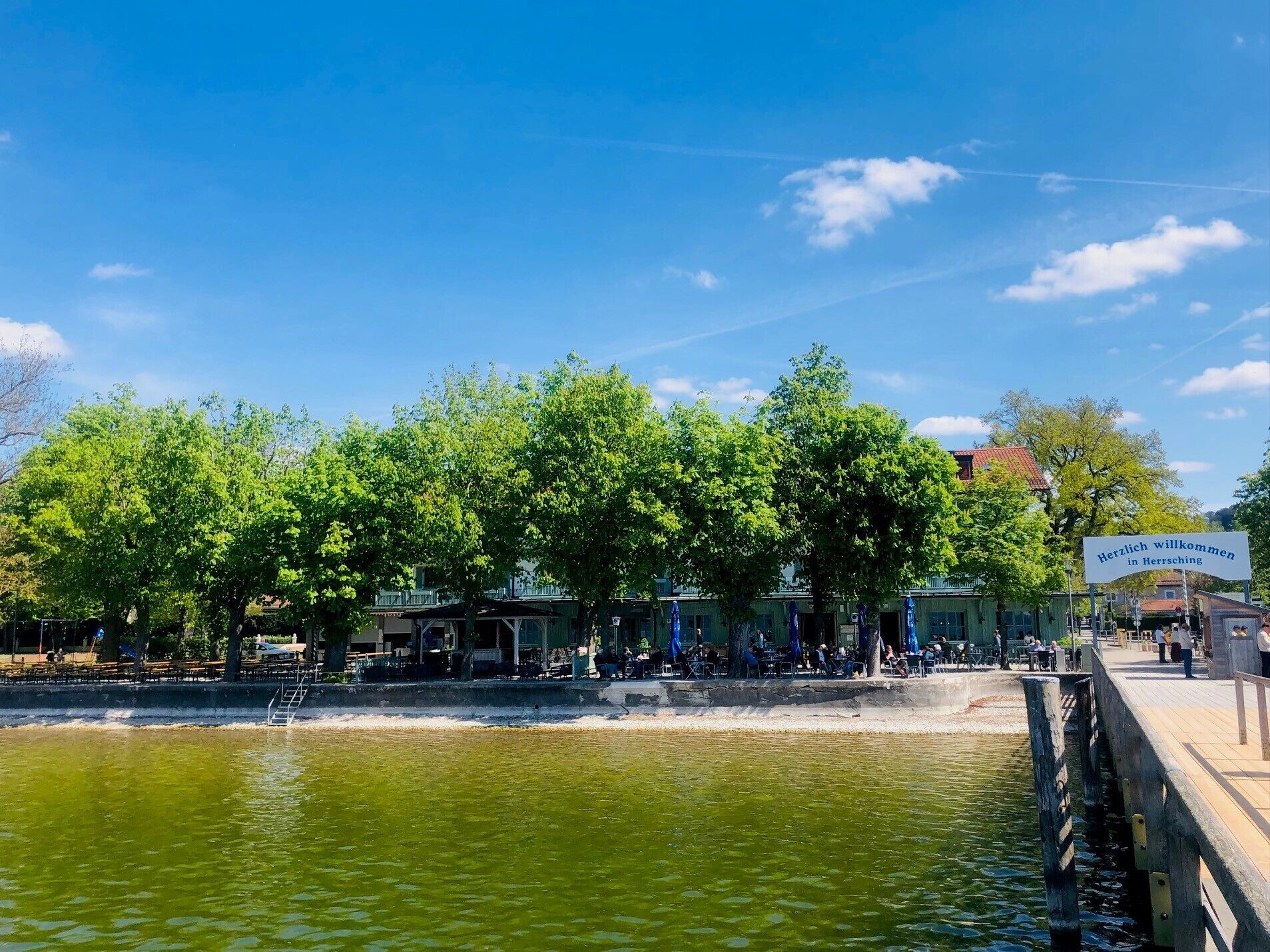 A riverside area with a wooden pier, trees, and a sign welcoming visitors to Herrsching.