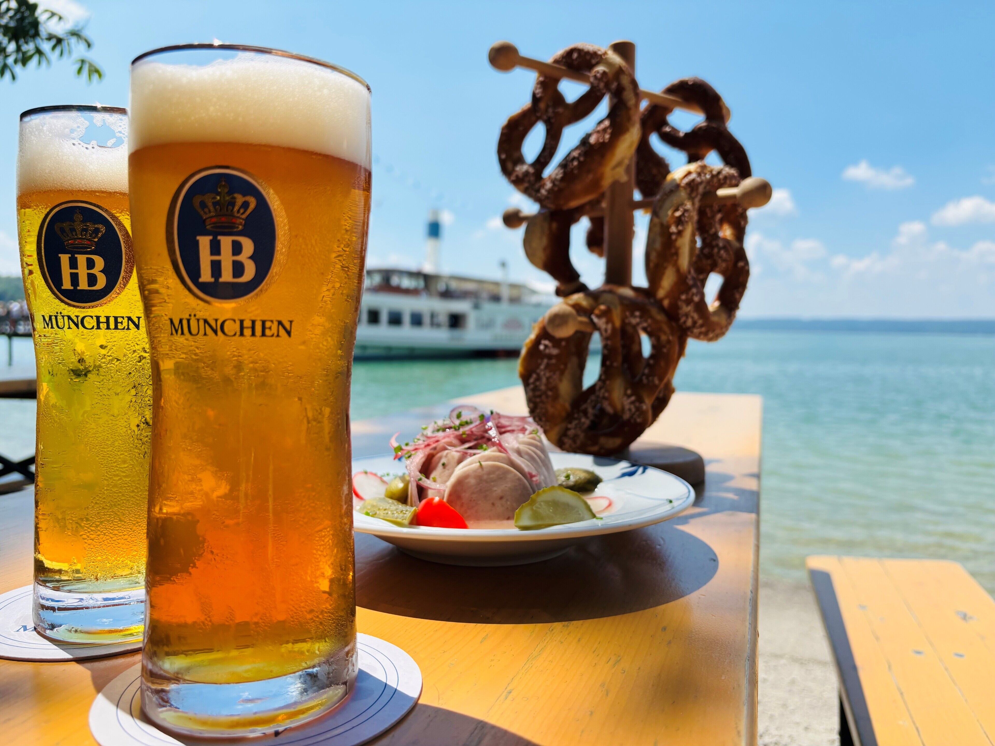 Two glasses of beer with 'München' branding, a plate of food, and a pretzel on a table by a waterfront.