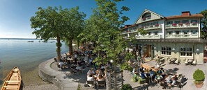 Un restaurant situé au bord du lac, avec une terrasse, entouré d’arbres et adossé à un immeuble doté de balcons.