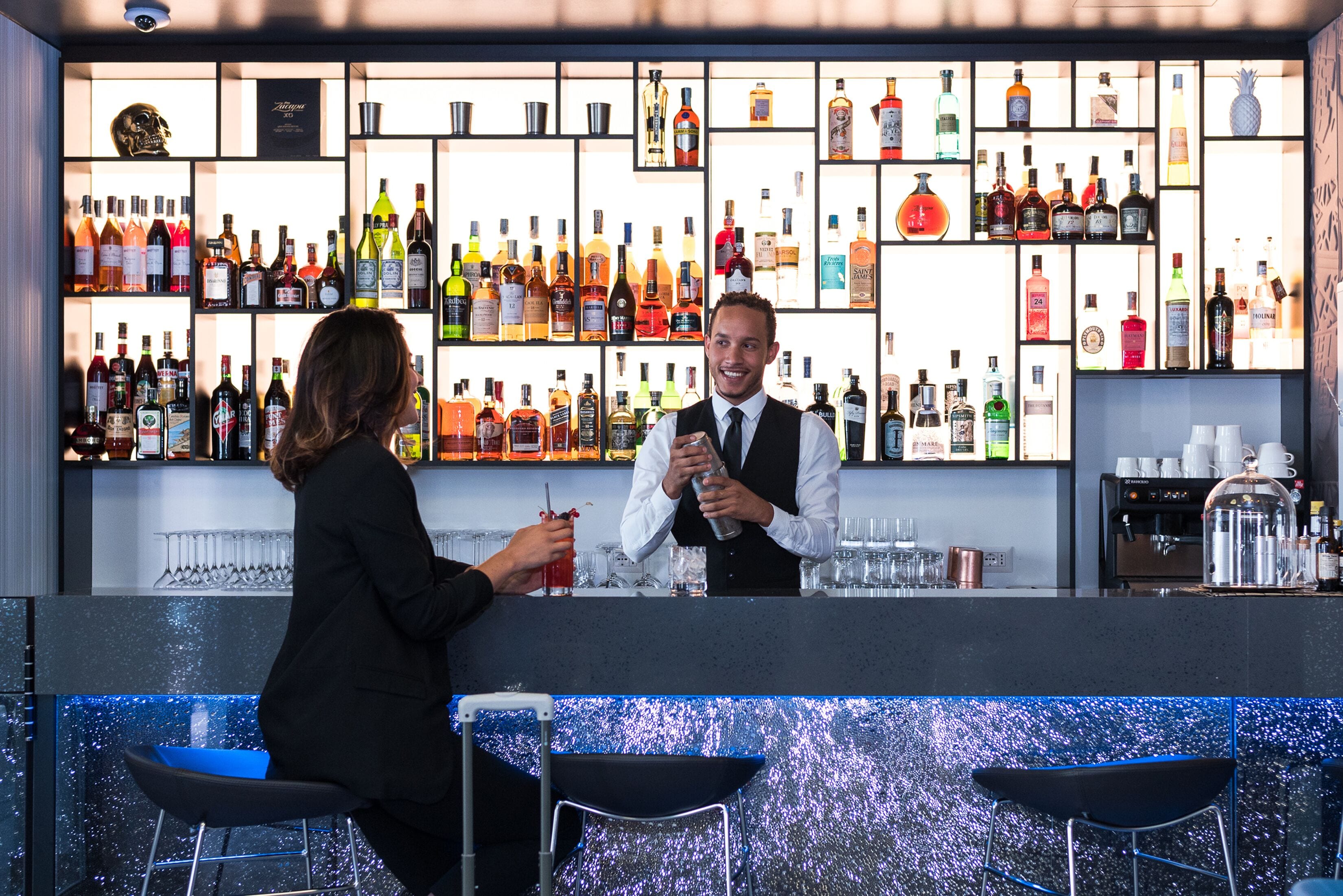 A bar area with a bartender and a customer, shelves of various liquor bottles, and bar stools.