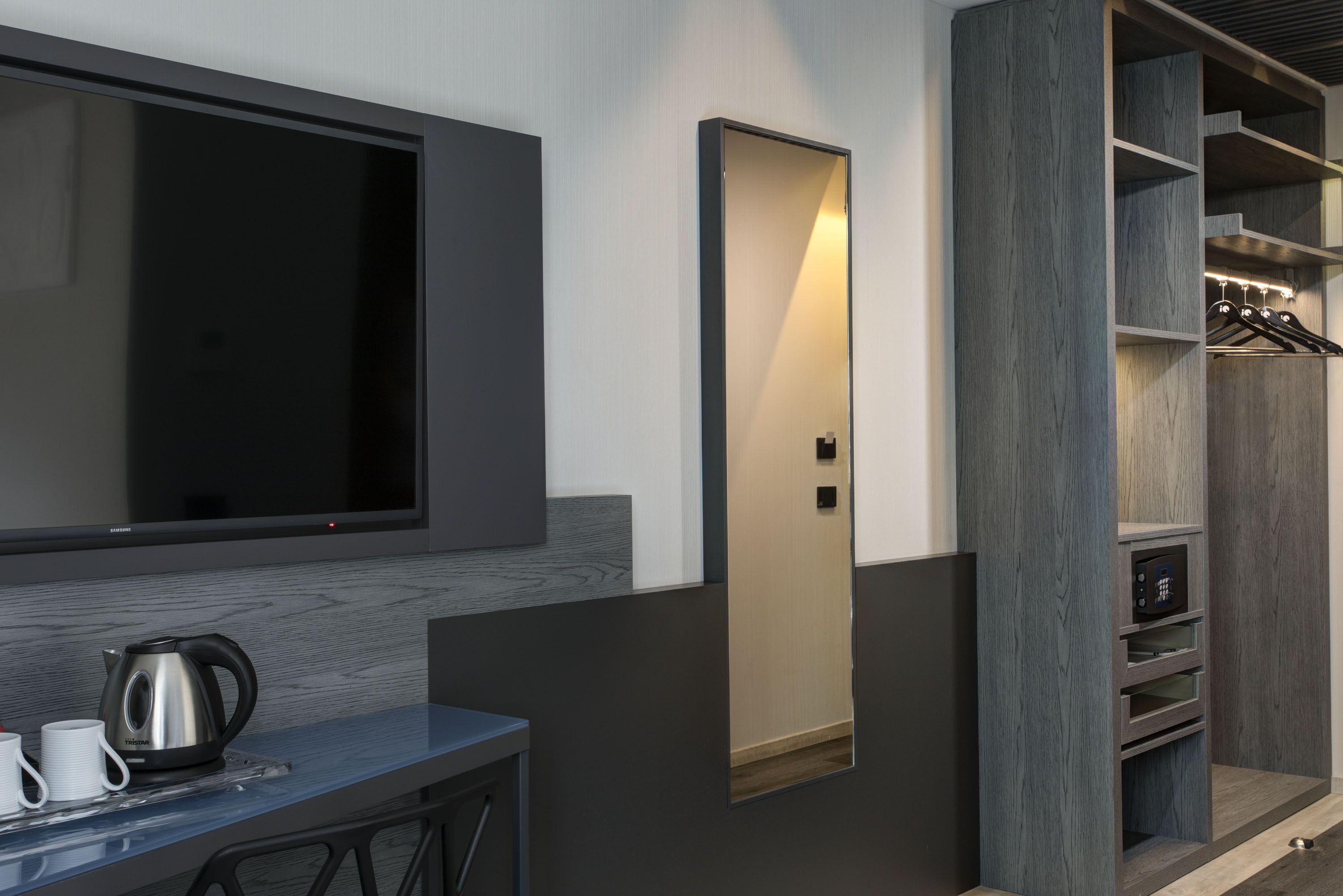Una habitación de hotel moderna con una televisión de pantalla plana montada en la pared, una mesita con una tetera y tazas, y un armario empotrado con espejo y repisas.