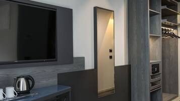 Una habitación de hotel moderna con una televisión de pantalla plana montada en la pared, una mesita con una tetera y tazas, y un armario empotrado con espejo y repisas.